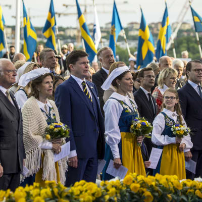 Kung Carl XVI Gustaf, drottning Silvia, riksdagens talman Andreas Norlén, kronprinsessan Victoria, prinsessan Estelle och prins Daniel firar Sveriges nationaldag på Skansen i Stockholm. Bakom dem vajar en rad svenska flaggor.