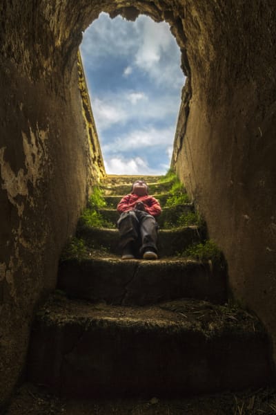 Ett barn sitter på en sliten mosstäckt trappa som går ner i ett mörkt rum. Barnet tittar upp mot den blå himlen.