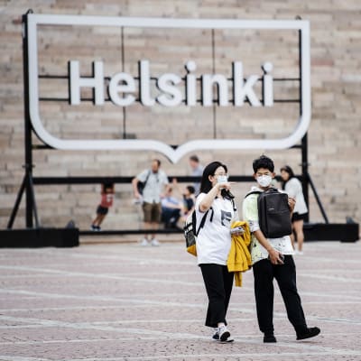 Ett par med munskydd går över Senaststorget i Helsingfors. I bakgrunden syns en stor skylt med texten "Helsinki".
