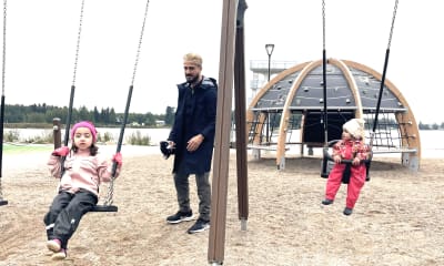 En man med svart jacka står och gungar två flickor i en lekpark