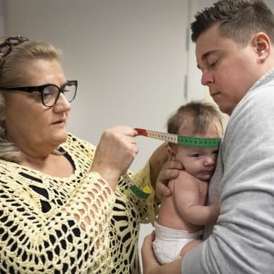 Terveydenhoitaja mittaa vauvan päänympärystä, vauva on isänsä sylissä.