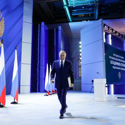 Vladimir Putin på väg bort från en talarstol. Han har ryska flaggor vid sin sida.