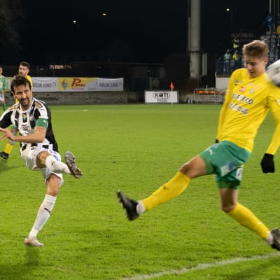 Fotbollsspelare skjuter ett hårt skott mot annan spelare på fotbollsplan under pågående match.