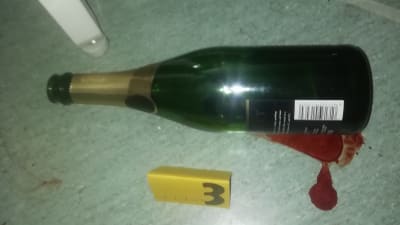En flaska för mousserande vin ligger i en blodpöl. Bredvid dem är en gul brottsutredningslapp med siffran tre.
