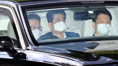 Tre japanska män sitter i en bil och de har munskydd på sig. En av männen är Japans premiärminister Shinzo Abe.