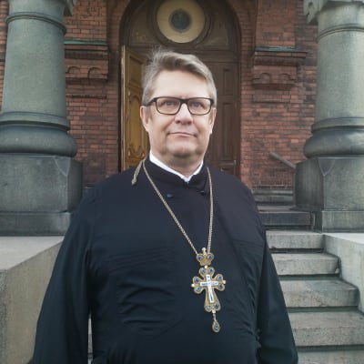 Markku Salminen, kyrkoherde vid Helsingfors ortodoxa församling, står framför en trappa in till Uspenskijkatedralen på Skatudden i Helsingfors. Han ser allvarsam ut och blickar ut bakom kameran. 