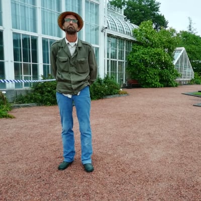 En man i hatt och mossgrön jacka står i Botaniska trädgården i Kajsaniemi i Helsingfors. Växthuset i bakgrunden.