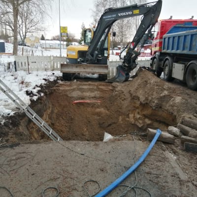 En stor och djup grop mitt i en väg i centrum av Ingå. Vattenledning som gått sönder. Grävmaskin och lastbil på plats. En lekpark till vänster om vägen.