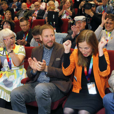 Li Andersson i orange jacka jublar med händerna i knytnävar. Paavi Arhinmäki sitter bredvid henne och applåderar tillsammans med andra partimedlemmar i bakgrunden.