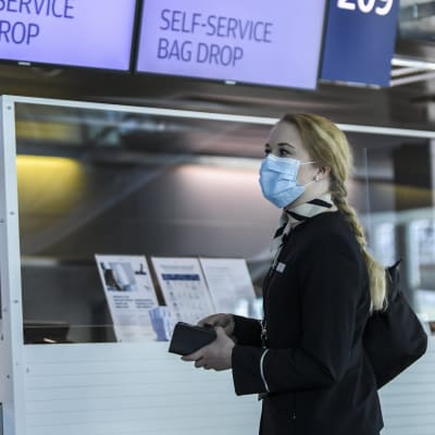 En kvinna med ansiktsskydd och Finnairs uniform står framför en check-in-disk.