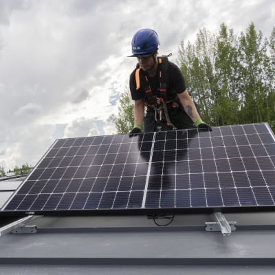 Aurinkopaaneelien asentaja Juha Matilainen asettelee suurta aurinkopaneelia kiskoille katolla.