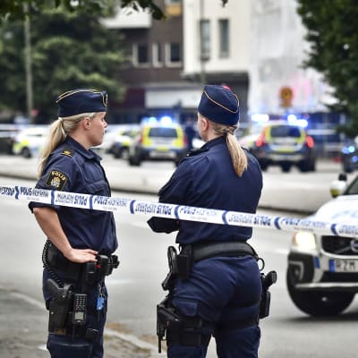 Två kvinnliga poliser bevakar ett avspärrat område. I bakgrunden står flera polisbilar.