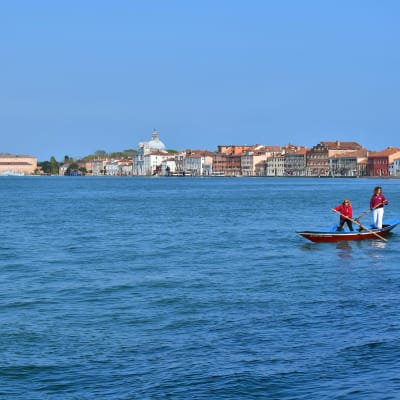 Två kvinnor står i en båt och paddlar sig fram på ett azurblått hav med Vendig i silhuett i bakgrunden.