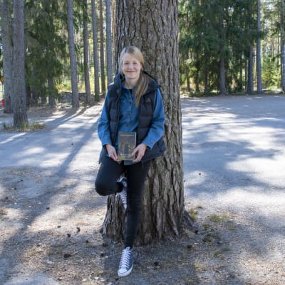 Mirkka Lappalainen lutar mot ett träd och håller i famnen sin bok Smittenin murha.