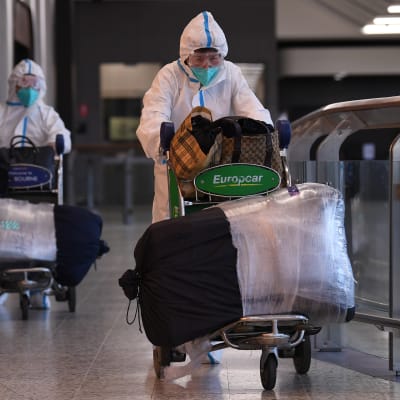 Kaksi suojapukuun pukeutunutta matkustajaa kävelee matkatavarakärryn kanssa Melbournen lentoasemalla. 