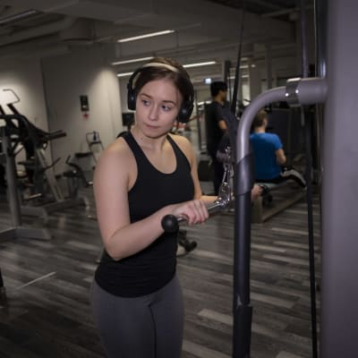 Nuori nainen treenaamassa kuntosalilla. Taustalla näkyy kuntosalilaitteita ja kaksi muuta treenaajaa. 