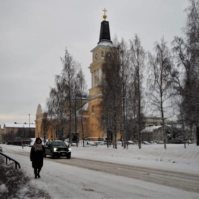 Talvinen katunäkymä, jossa nainen kävelee jalkakäytävällä ja tumma auto ajaa kadulla. Taustalla koivuja ja keltainen iso Oulun tuomiokirkko.
