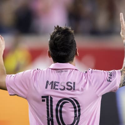 Lionel Messi fotograferad bakifrån, gör en målgest med pekfingrarna i luften, iklädd en ljusröd skjorta med nummer 10 på ryggen. 