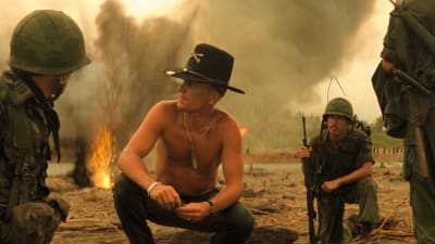 Bill Kilgore (Robert Ruvall) sitter på huk på en sandstrand omgiven av soldater och explosioner.