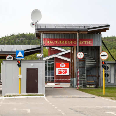 En mindre gränsstation med nordliga fjäll i bakgrunden. Skyltarna som syns är på ryska och engelska. Bakom gränsövergången skymtar en taxfree-butik.