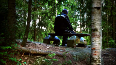 Man sitter ensam och bortvänd på bänk i skogen.