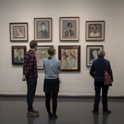 Helene Schjerfbeck-utställning på Ateneum. Tre personer med ryggen mot kameran kollar på tavlor.