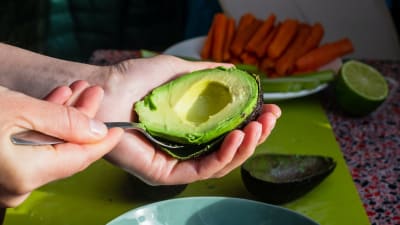 Någon skalar en avokado, en halva ligger i en turkos skål 