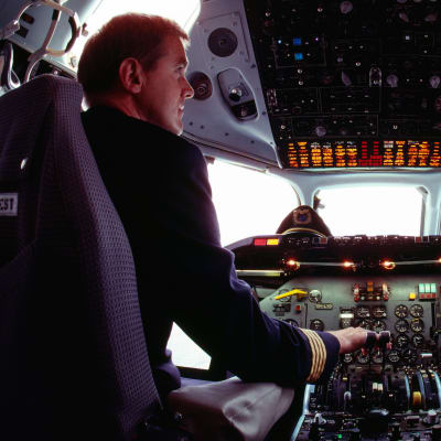 Två piloter sitter i ett flygplan vid styrpanelen. De tittar på varandra medan de sitter på varsin stol.