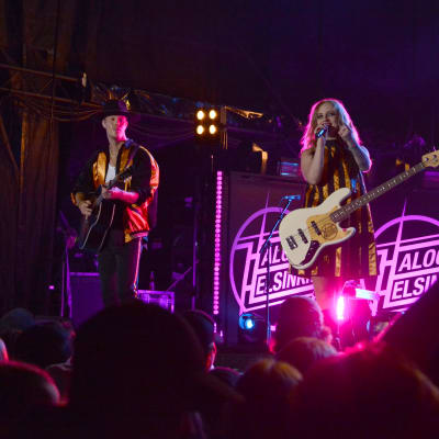 Rock-yhtye esiintyy lavalla, taustalla näkyy laulaja-basisti Elli Haloo (Elisa Tiilikainen) ja kitaristi Jere Marttila