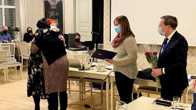 Flera personer tittar på två kvinnor som kramas. De andra håller i blommor och ett häfte.
