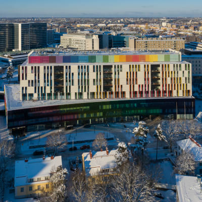 Uusi lastensairaala Helsingissä ilmasta kuvattuna. On talvi, maassa on lunta ja aurinko paistaa kirkkaasti. Lastensairaalan seinän yläosassa näkyy paljon eri värejä. 