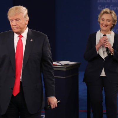 Trump seisoo etualalla, Clinton taempana. Trumpin ilme on vakava, Clinton hymyilee.