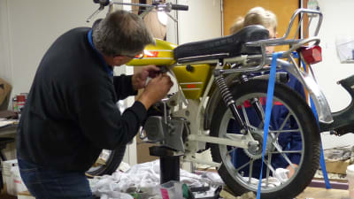 En man reparerar en mooped som står på ett bord i en verkstad. Bakom mopeden kan man skymta några unga pojkar.