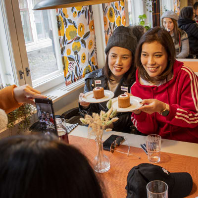 Två thailändska kvinnor sitter i ett café. De poserar med två Runebergstårtor på ett fat och en kvinna som sitter mitt emot tar bilden med sin telefon.