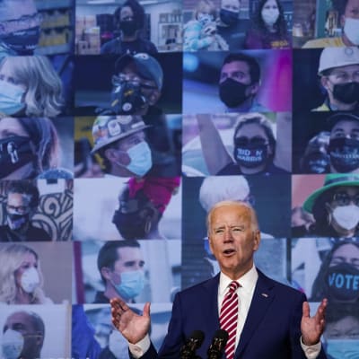 Joe Biden vid mikrofoner, framför en bildvägg med angängare iklädda coronaskydd.