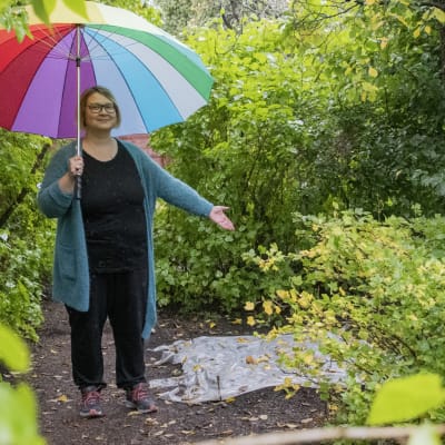 En kvinna med ett färgglatt paraply står i en trädgård och pekar ut ett område med en presenning på.