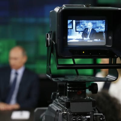 Presidentti Vladimir Putin näkyy häilyvänä kameran taustalla, RT-kanavan kuvaukset käynnissä.