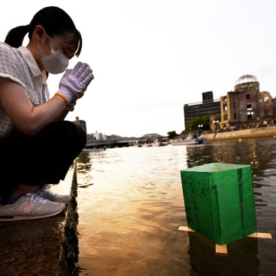 En person sitter på huk vid en vattenlykta. I bakgrunden syns minnesmärket för Hiroshimas bombning.