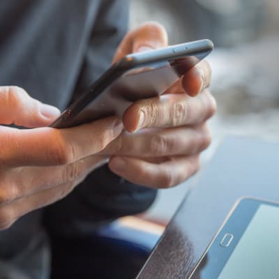 Två händer som knapprar på en svart smarttelefon. I den högra kanten syns en surfplatta.