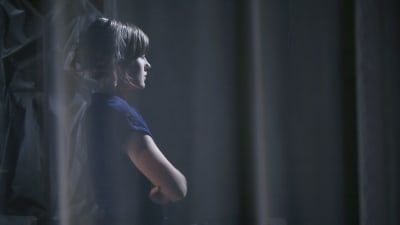 En ung kvinna står i profil mot en mörk bakgrund bakom en genomskinlig gardin.