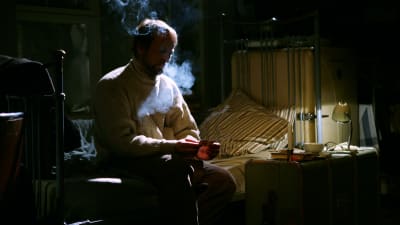 En man i ljus tröja sitter på en säng och röker.