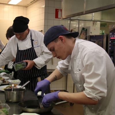 Två unga män i förda kockkläder och handskar lägger upp mat på fat i ett skolkök