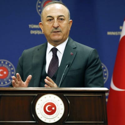 Mevlüt Çavuşoğlu står vid ett talarpodium och pratar samtidigt som han gestikulerar med händerna. Han är klädd i mörk kostym med slips.
