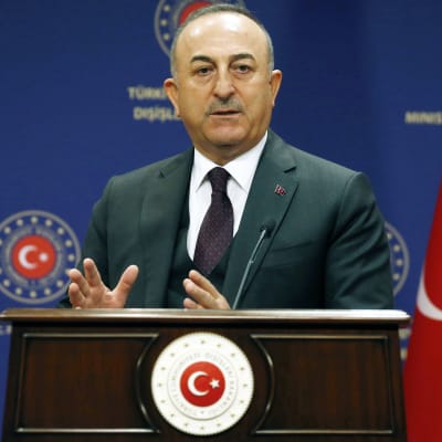Mevlüt Çavuşoğlu står vid ett talarpodium och pratar samtidigt som han gestikulerar med händerna. Han är klädd i mörk kostym med slips.
