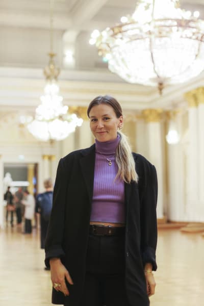 Märta Westerlund iklädd i kostymbyxor, kavaj och lila topp står i en sal i presidentens slott. Hon ler och tittar in i kameran.