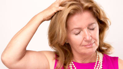 Kvinna med pärlhalsband håller sig för huvudet och blundar