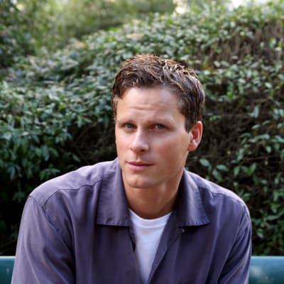 Skådespelaren Adam Pålsson sitter på en bänk i ett grönområde och tittar in i kameran. Han har en ljusblå tröja på sig.