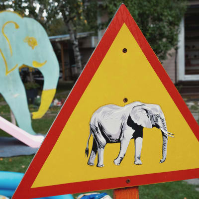 Varoituskolmio, jossa norsun kuva. Taustalla lasten leikkipaikka.