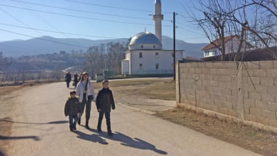 Två pojkar och en kvinna går på en väg. I bakgrunden syns en moské.