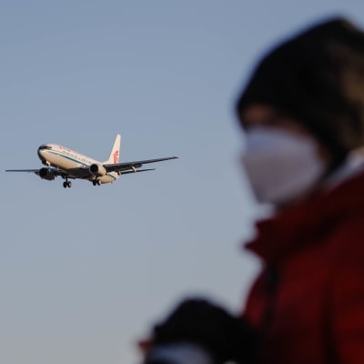 Ett flygplan avtecknar sig mot himlen. I förgrunden står en oskarp figur, men det är tydligt att personen bär munskydd, vintermössa och jacka.